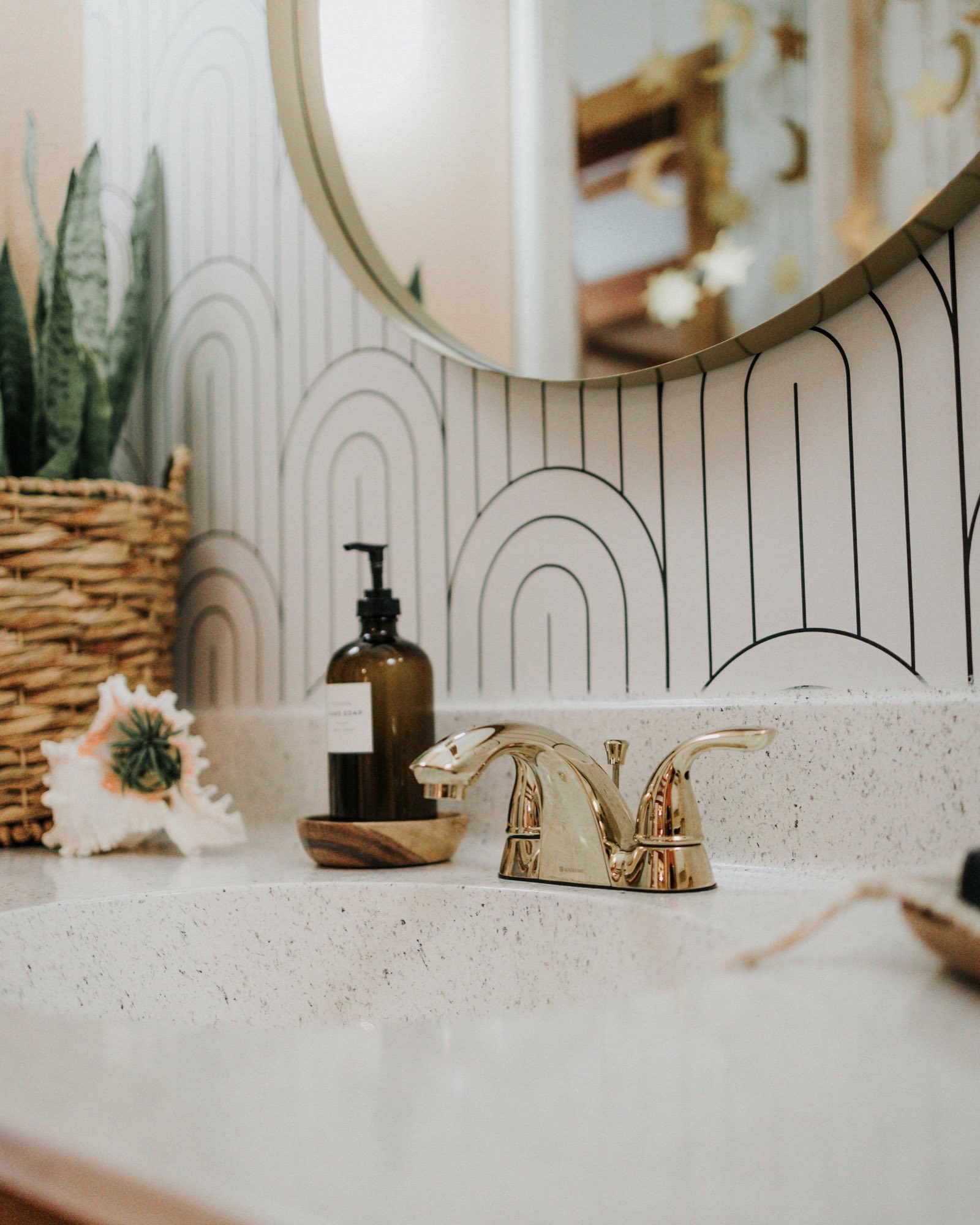Close view of a bathroom vanity with a polished brass faucet, terrazzo counter, and a round brass mirror against arched wallpaper