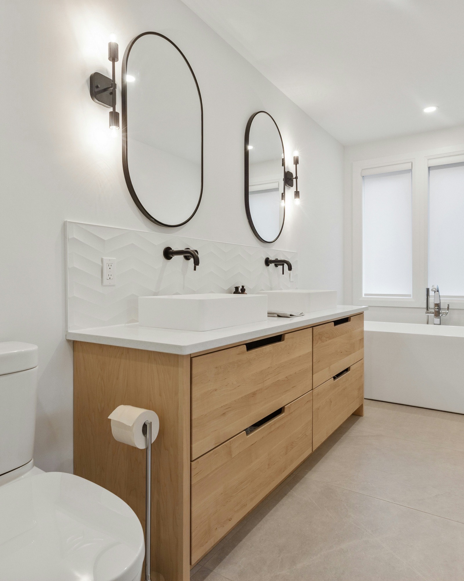 Bathroom with a white oak floating double vanity, vessel sinks, round black mirrors, and a freestanding tub