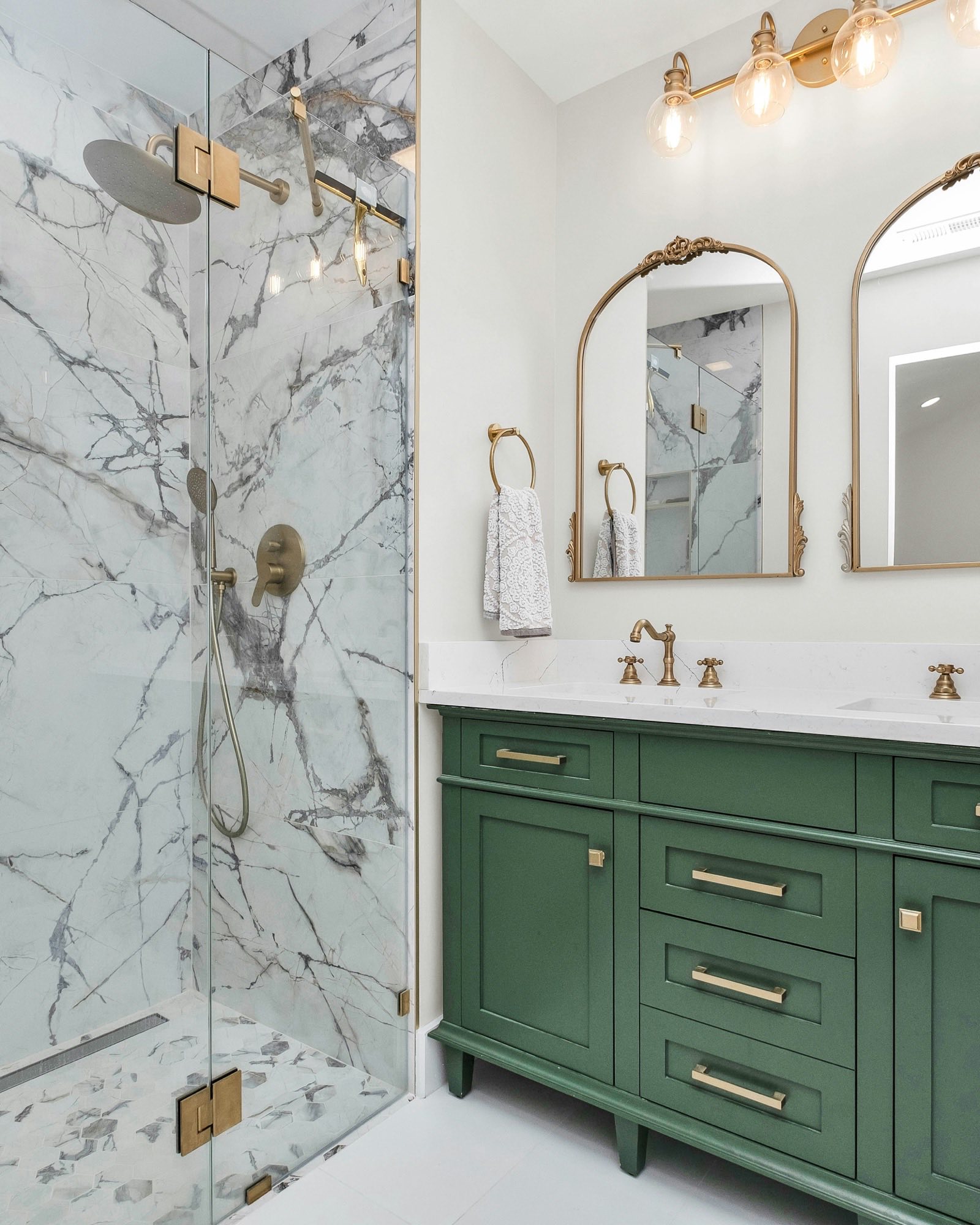 Walk-in shower wrapped in marble slab beside a green double vanity with brass fixtures and arched mirrors