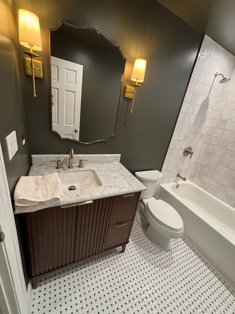 Bathroom with dark walls, marble-topped wood vanity, brass sconces, and basketweave tile floor